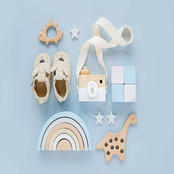 Assortment of baby essentials including shoes, wooden toys, camera, and rainbow stacker on blue background.