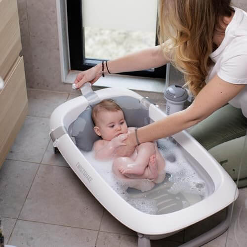 Mother gently bathing her baby in a modern Twistshake baby bathtub near a window.
