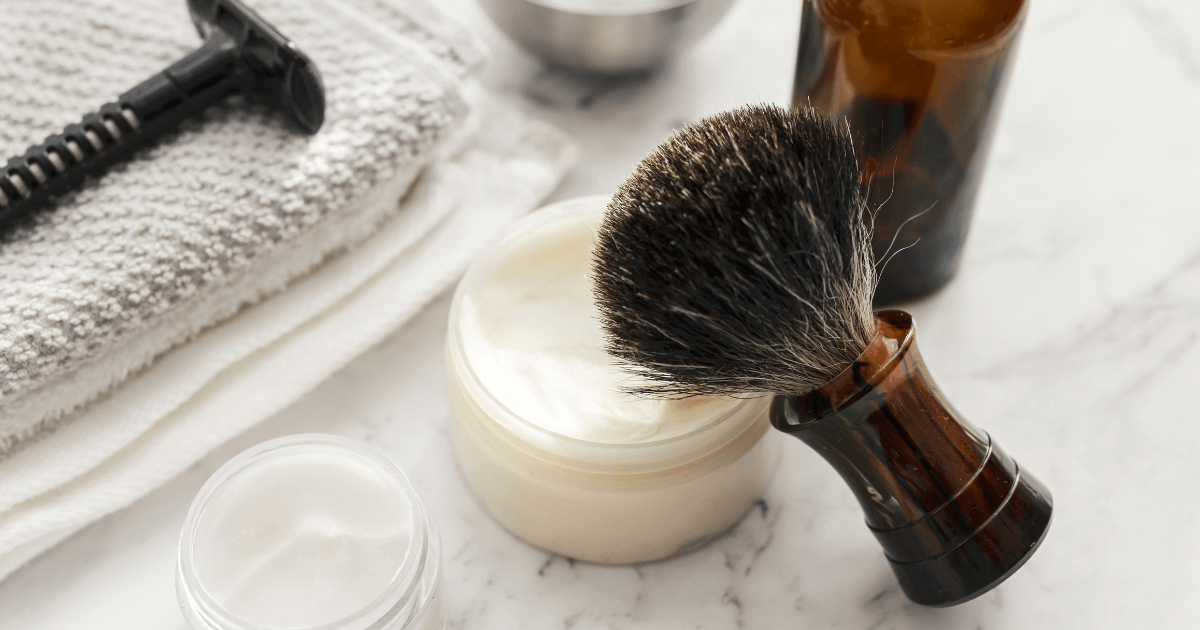 Assorted beard grooming tools including a shaving brush, cream, and trimmer on a marble surface.