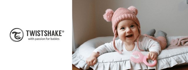 Joyful baby in pink hat holding Twistshake teether, lying in a baby nursery for Twistshake Baby Accessories ad.